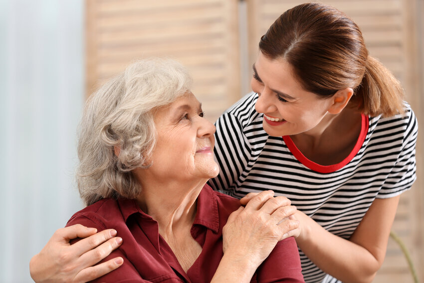 Elderly woman with grey hair sitting and wearing a burgundy blouse, being supported and comforted by a younger caregiver in a striped shirt; scene suggests caregiving and emotional support for Alzheimer’s disease — Ace QBank, Clinical Edge.
