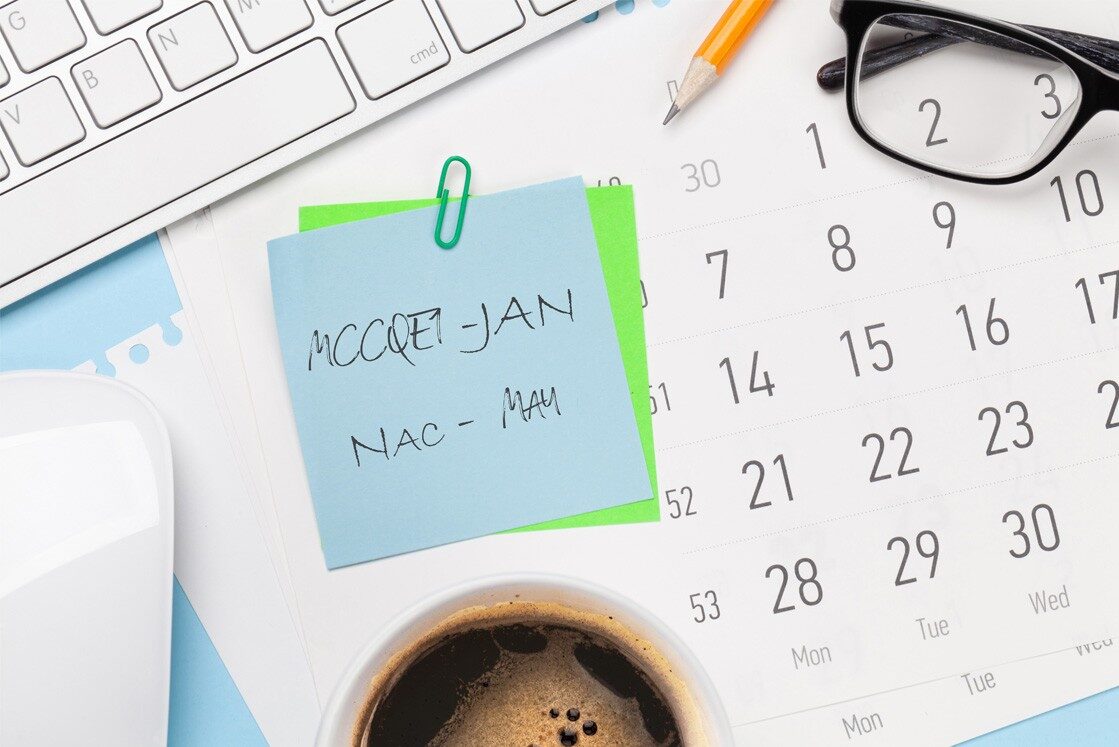 A calendar, sticky note with handwritten reminders, keyboard, glasses, pencil, mouse, and a cup of coffee on a desk. Perfect for MCCQE1 exam preparation and tracking important 2026 MCCQE1 Exam Dates.