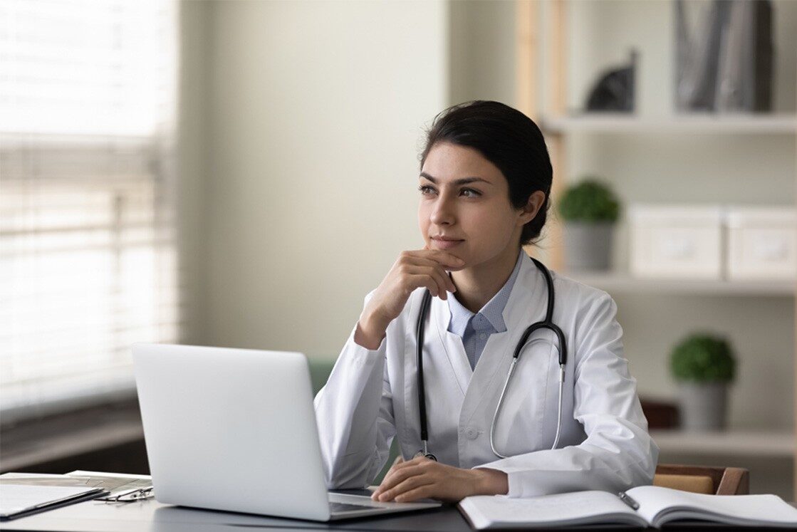 A doctor wearing a white coat and stethoscope sits at a desk with a laptop and open notebook, looking thoughtfully to the side. Perhaps contemplating MCCQE1 exam preparation or what MCCQE1 will actually demand in 2026.