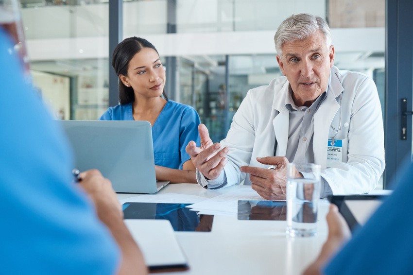 A doctor wearing a white coat and stethoscope sits at a desk with a laptop and open notebook, looking thoughtfully to the side. Perhaps contemplating MCCQE1 exam preparation or what MCCQE1 will actually demand in 2026.