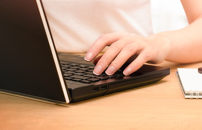 A person types on a black laptop with their right hand while sitting at a wooden desk, perhaps researching how to achieve the best MCCQE1 result. A notepad and pen are partially visible beside the laptop.