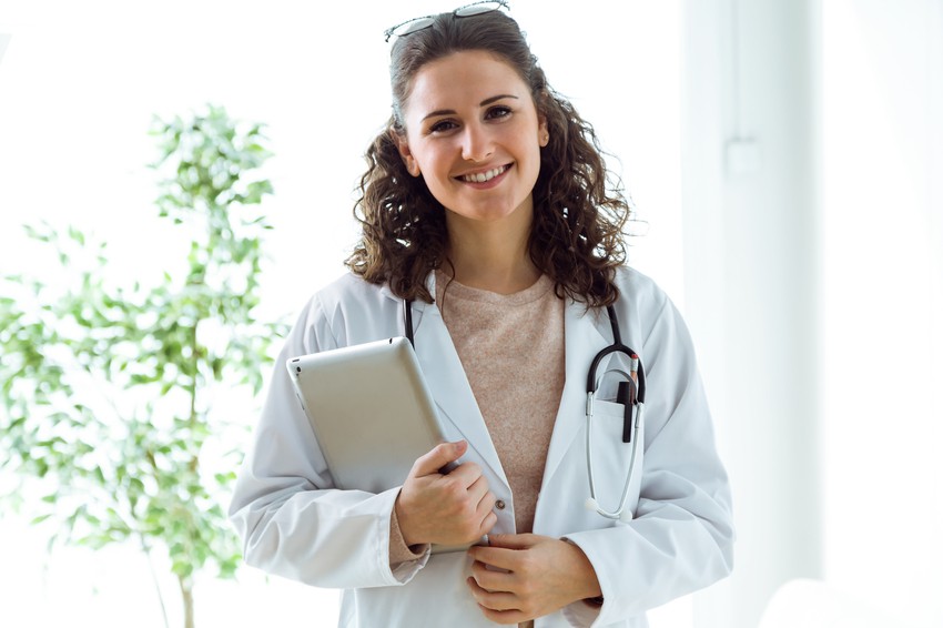A woman in a white lab coat holds a tablet and smiles at the camera, with a stethoscope draped over her shoulder. Ready to share tips on how to achieve the best MCCQE1 result.