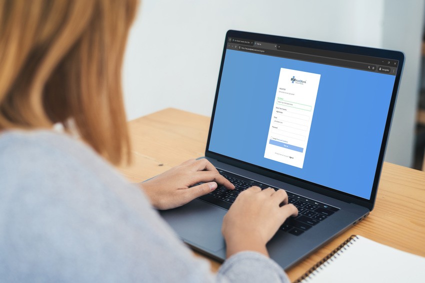 A person uses a laptop to fill out an online registration form on a website, with a notebook beside them on the wooden desk to note important MCCQE1 exam dates from Ace QBank.