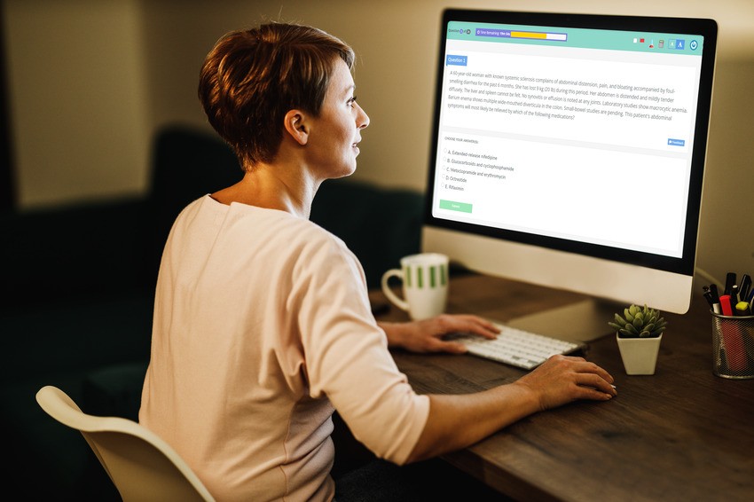A person is sitting at a desk using a desktop computer, reading MCCQE1 exam dates displayed on the monitor in a home office setting, likely while preparing with Ace QBank.