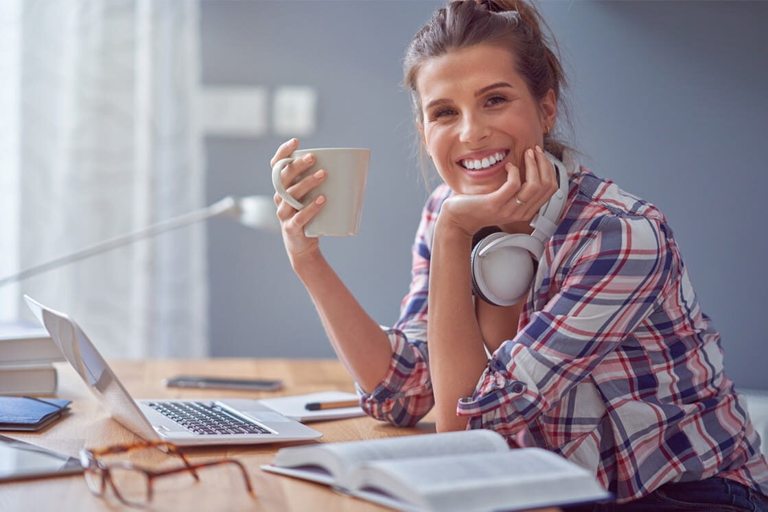 A woman wearing headphones around her neck sits at a desk with a laptop, open book and mug, smiling confidently at the camera. Ready for the April 2026 MCCQE1 Exam with Ace QBank by her side.