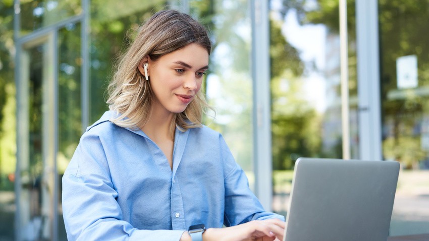 A woman wearing earbuds and a blue shirt works on a laptop outdoors, preparing for MCCQE1 with Ace QBank. Surrounded by glass windows and greenery.