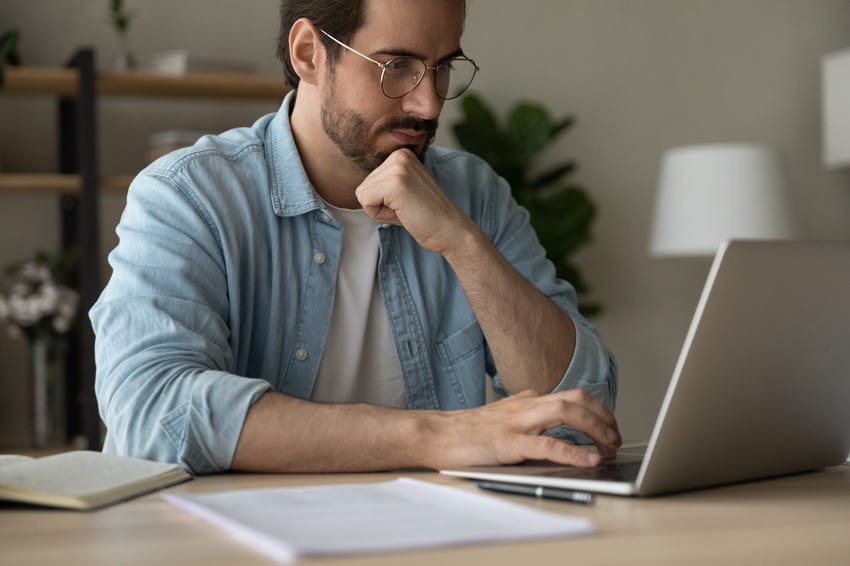 Man wearing glasses and a denim shirt sits at a desk. Looking thoughtfully at his laptop screen as he reviews MCC Objectives and Ace QBank MCCQE1 practice questions, with a notebook and papers nearby.