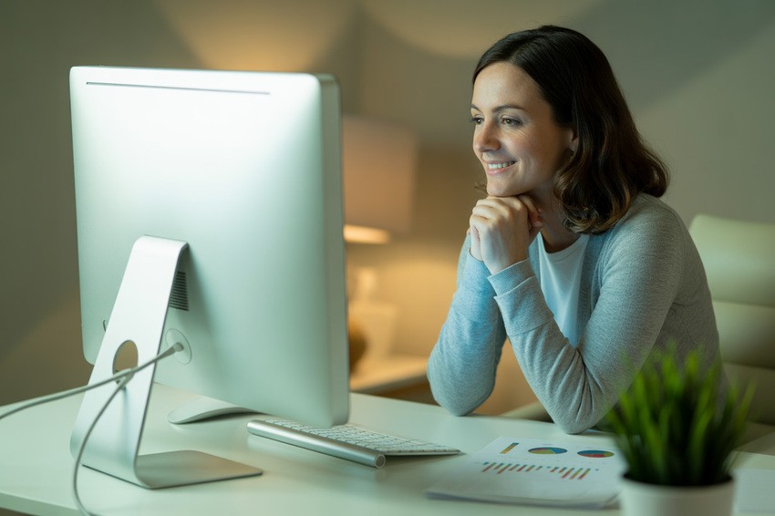 A woman sits at a desk, smiling at a large computer monitor. Papers with charts, MCC Objectives & MCCQE1 Practice Questions, are on the desk next to a keyboard and a potted plant. Perfect for focused MCCQE1 exam preparation with Ace QBank.