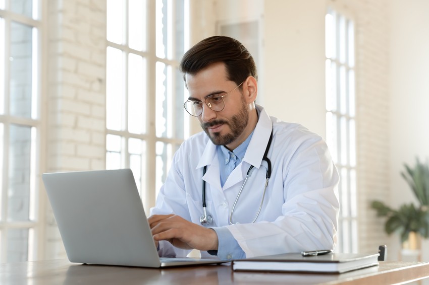 A doctor wearing a white coat and stethoscope works on a laptop at a desk in a bright office, checking Ace QBank to prepare for the MCCQE1 exam dates and updates on the 2026 MCCQE1 Scheduling Open for April.