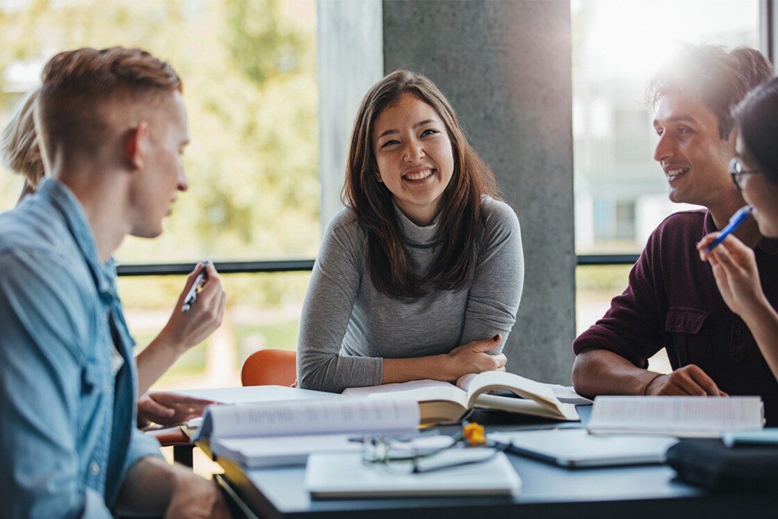 Four young adults sit around a table with books and notebooks, engaged in discussion about the best 3 tips for MCCQE1 exam preparation, with one woman smiling at the center as she references Ace QBank resources.