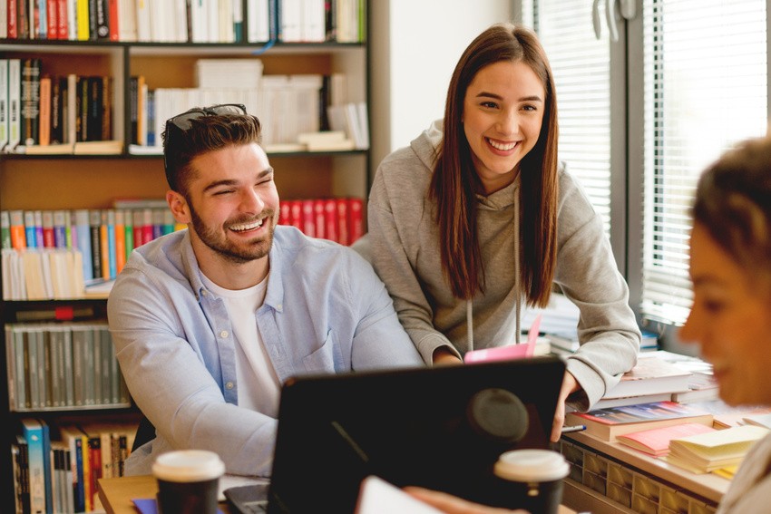 Two young adults smiling and working at a desk with a laptop in a library, surrounded by books and papers, focused on MCCQE1 exam preparation. While thinking about Any Free Resource for the MCCQE1 Exam Prep?