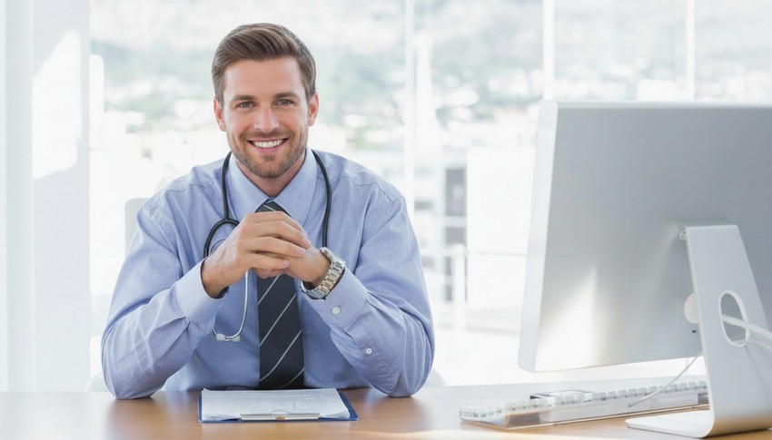 A man in a dress shirt and tie, with a stethoscope around his neck, sits at a desk in an office, smiling at the camera. Ready for MCCQE1 Exam Preparation with Ace QBank.