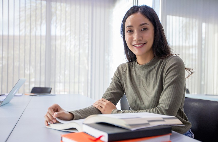 A woman sits at a table in a bright room, smiling at the camera with open books and notebooks. Ready to master the MCCQE1 using Ace QBank, learning what top candidates do differently.