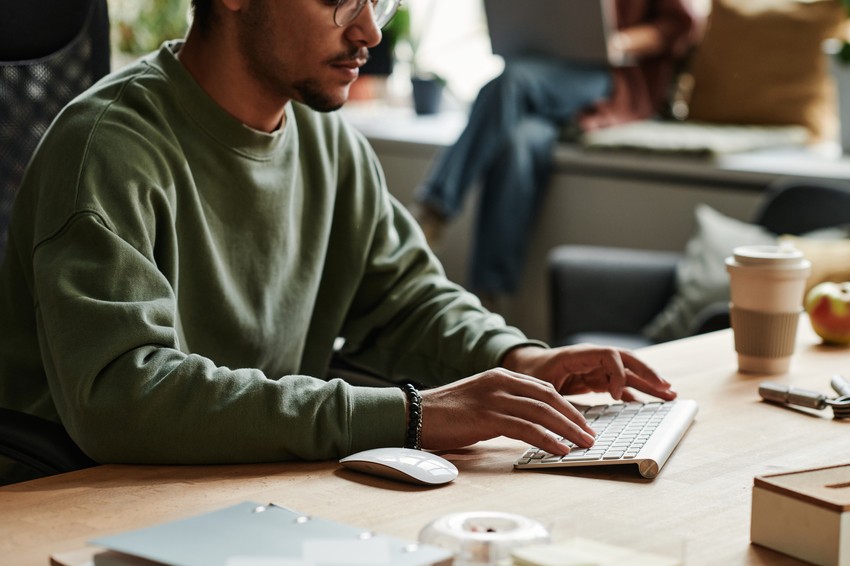 A person in a green sweatshirt types on a wireless keyboard at a wooden desk, perhaps researching why most MCCQE1 study plans fail, with a coffee cup, papers, and office items nearby. Ace QBank materials may be present.