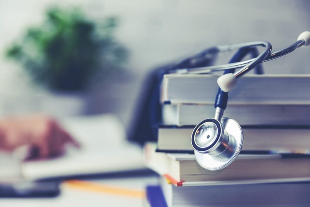 A stethoscope rests on a stack of books on a desk, with a blurred hand writing in the background and a potted plant to the side. Capturing the focused dedication of candidates preparing for the MCCQE exam. This scene reflects the spirit behind Ace QBank as medical professionals gear up for the Countdown to August-September 2026 MCCQE Exam Dates.