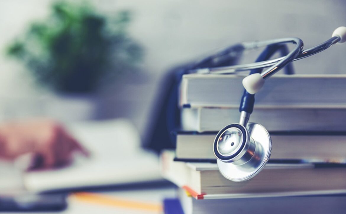 A stethoscope rests on a stack of books on a desk, with a blurred hand writing in the background and a potted plant to the side. Capturing the focused dedication of candidates preparing for the MCCQE exam. This scene reflects the spirit behind Ace QBank as medical professionals gear up for the Countdown to August-September 2026 MCCQE Exam Dates.