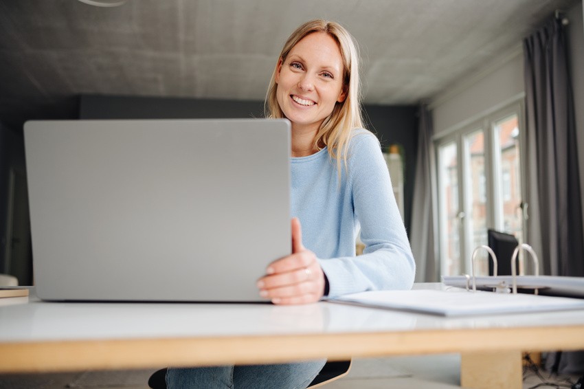 Woman sitting at a desk, smiling at the camera, with an open laptop in front of her in a bright, modern room—she’s ready to monitor MCCQE1 exam preparation and review demo questions for success.