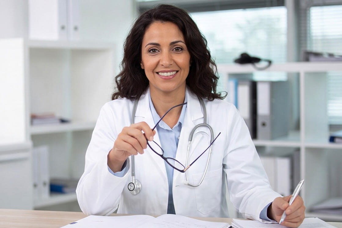 A woman doctor in a white coat sits at a desk, holding glasses in one hand and a pen in the other, smiling at the camera. Ready to share expert tips on how to improve your time for the MCCQE with resources like Ace QBank.