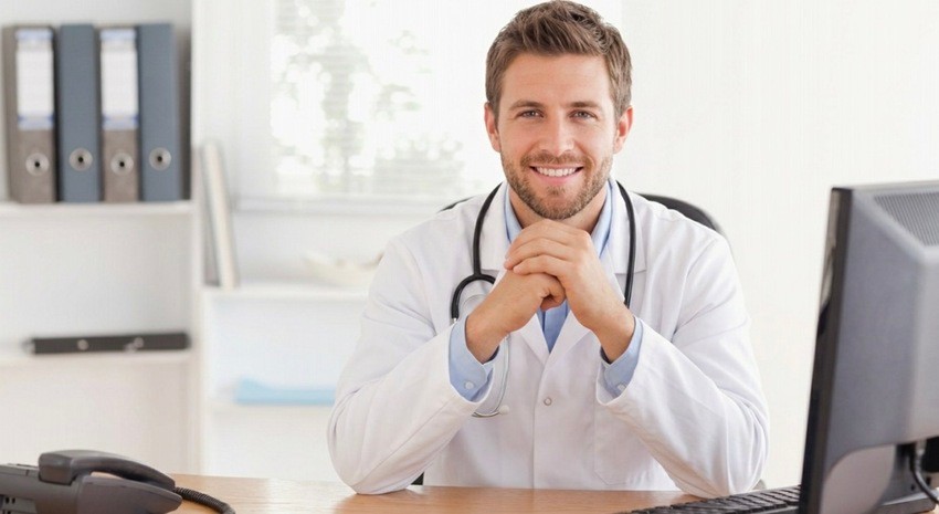 A male doctor in a white coat sits at a desk, smiling, with his hands clasped in front of a computer and a telephone. Perhaps reflecting on how to improve your time for the MCCQE using resources like Ace QBank.