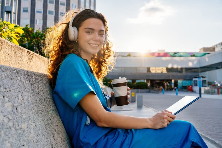 Woman in medical scrubs sits outside, holding a tablet and a coffee cup, wearing headphones, with sunlight in the background. Preparing for the Countdown to August-September 2026 MCCQE Exam Dates using Ace QBank.