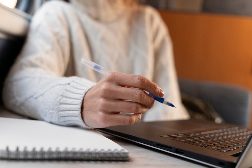 A person in a white sweater holds a pen over a laptop keyboard, with an open notebook on the table nearby. Leveraging colour highlights for MCCQE1 exam prep with Ace QBank.
