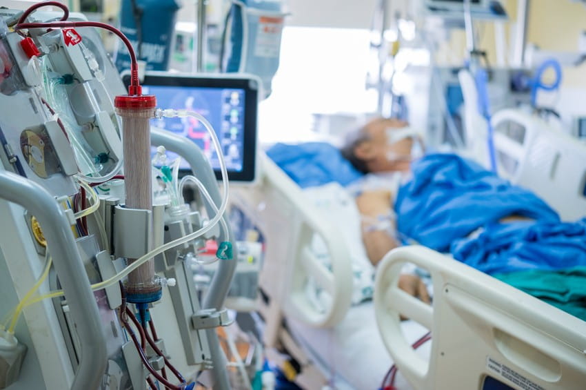 A hemodialysis machine is in the foreground, with a blurred patient resting in a hospital bed in the background. A scene that could be featured in MCCQE1 study cases or Ace QBank resources on topics such as Hantavirus-induced renal complications.