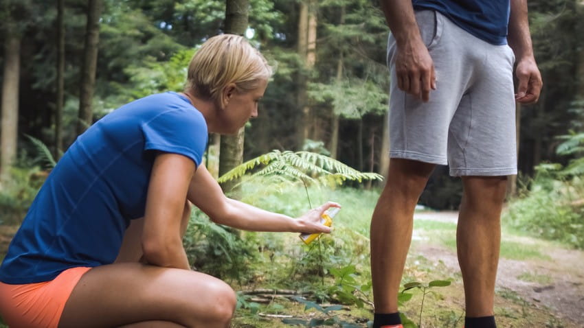 A person crouches in the woods, spraying insect repellent on another person’s legs to help protect against tick bites and Lyme Disease. Both are dressed in athletic clothes.