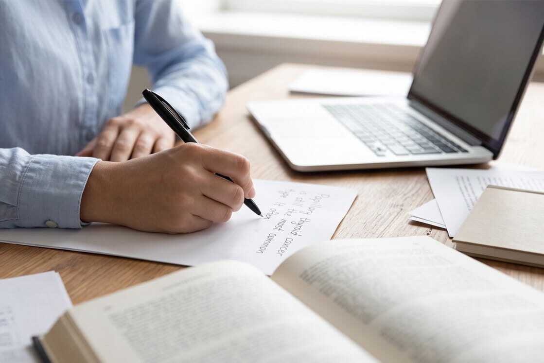 Person writing on paper at a desk with an open laptop, book, and documents, suggesting studying, researching, or working in an academic or professional setting. Perhaps illustrating Why Take Notes During MCCQE Question Practice with Ace QBank to enhance retention and success on the MCCQE.