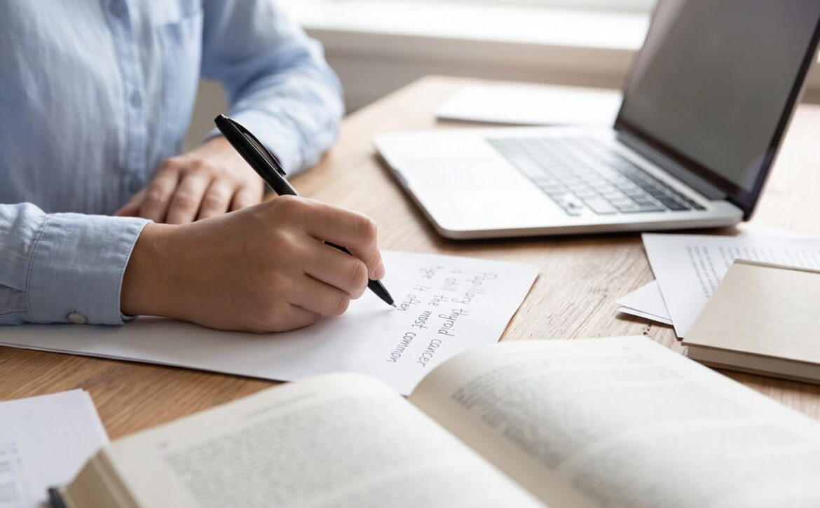Person writing on paper at a desk with an open laptop, book, and documents, suggesting studying, researching, or working in an academic or professional setting. Perhaps illustrating Why Take Notes During MCCQE Question Practice with Ace QBank to enhance retention and success on the MCCQE.